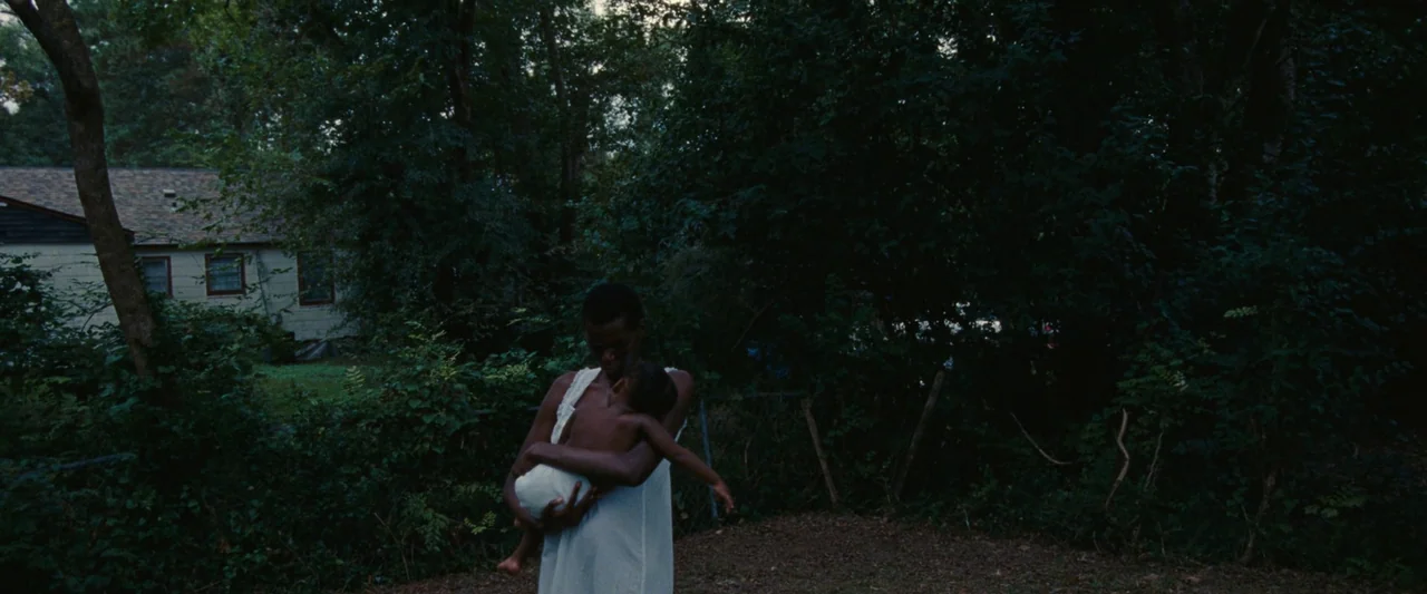 All Dirt Roads Taste of Salt (2023) movie still: medium — A backyard at dusk features dense foliage and a house visible in the background. A woman…