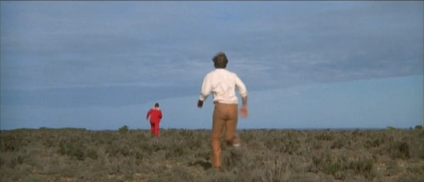 Roadgames (1981) movie still: wide — An open expanse of arid, scrubby land stretches to a distant horizon under a blue sky. Two men in casual…