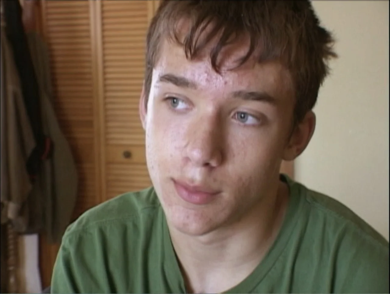 Addiction (2024) movie still: closeup — A domestic interior with a slatted wooden door visible in the background. A teenage boy sits facing the…