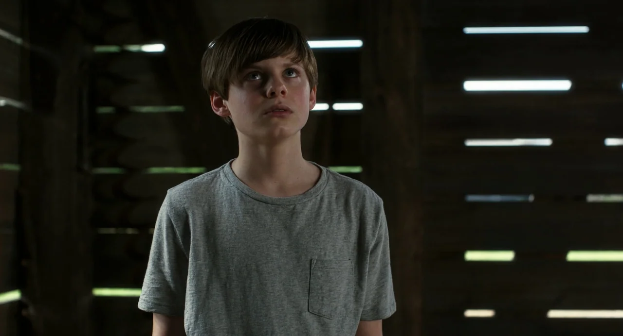 Goodnight Mommy (2022) movie still: medium — The interior of a dim barn with horizontal wooden planks allowing streaks of light. A young boy stands…
