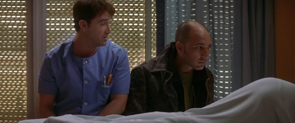 Talk To Her (2002) movie still: medium — A hospital room with a patient under a white sheet in the foreground. Two men sit behind the bed in front…