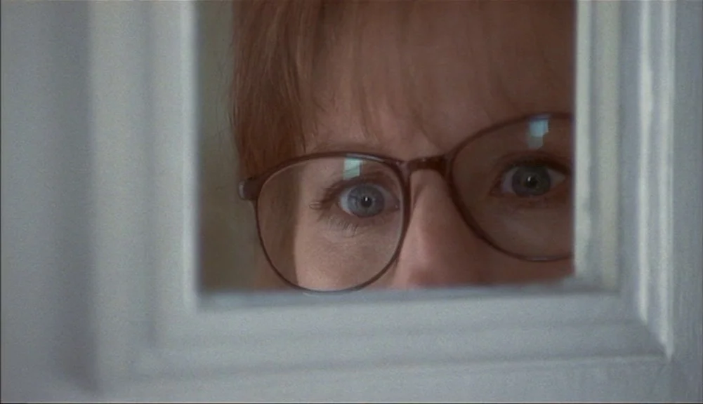 The Mirror Has Two Faces (1996) movie still: closeup — A woman looks through a rectangular glass windowpane set into a white door. Her face is…