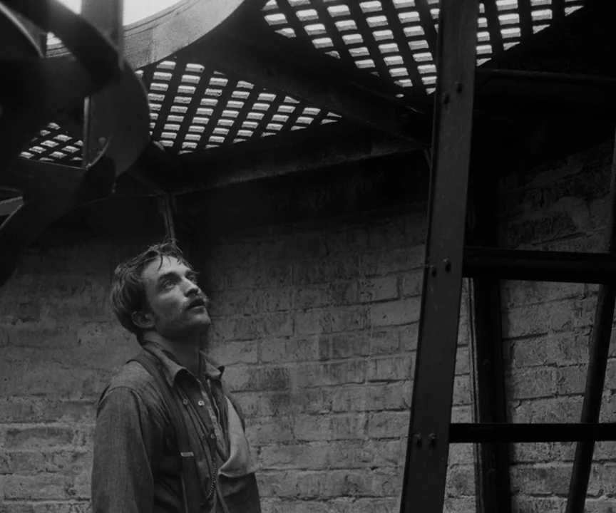 The Lighthouse (2019) movie still: medium — A man stands beneath a weathered outdoor iron staircase against a rough brick wall. He gazes upward…