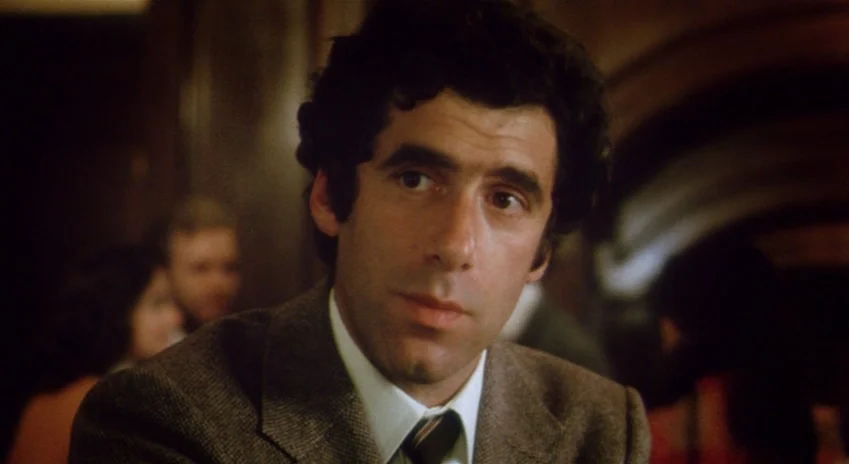 The Silent Partner (1978) movie still: closeup — A dimly lit indoor restaurant setting with blurred background figures. A man with curly hair looks…