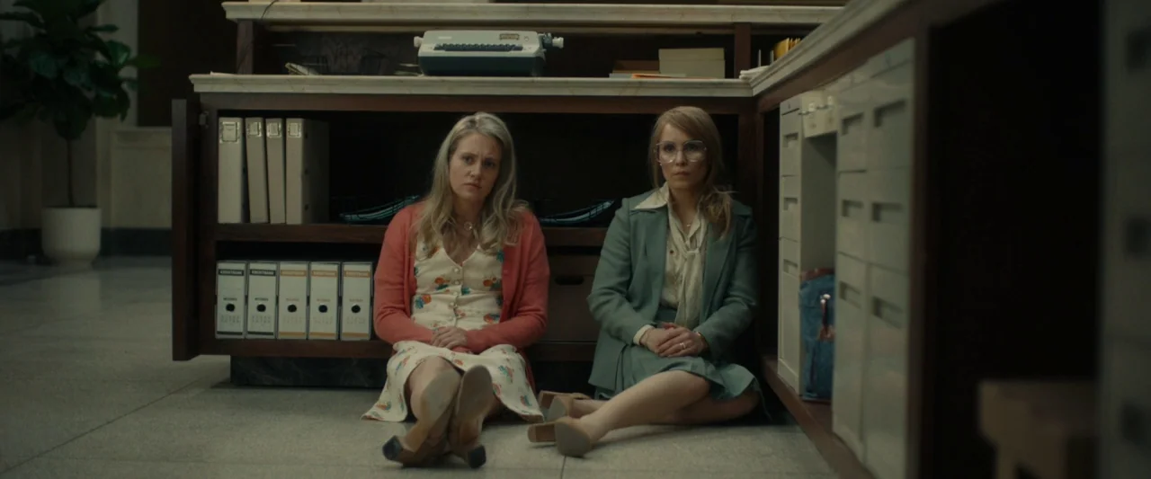 Stockholm (2019) movie still: fullbody — Two women sit on a tiled floor beneath a wooden counter in a bank. Files and a vintage typewriter rest on…