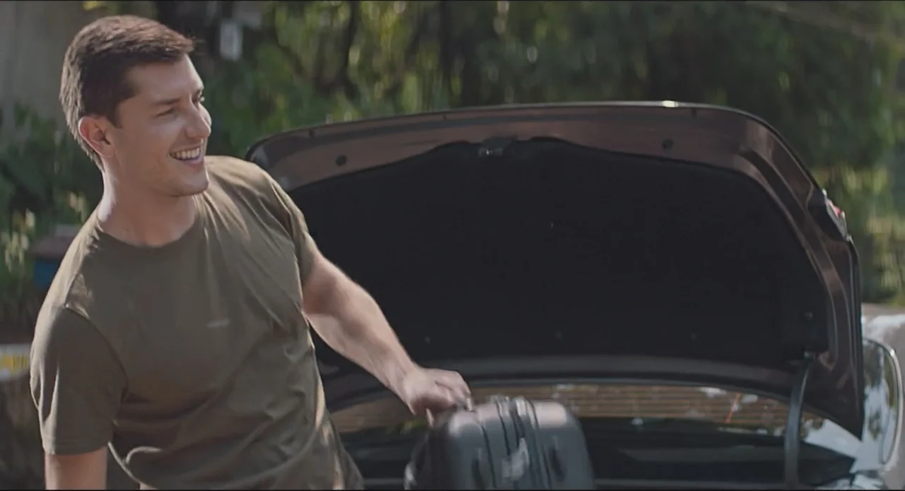 Proof of Love (2022) movie still: medium — A man stands next to an open car trunk outdoors with green foliage visible behind. He is placing a small…