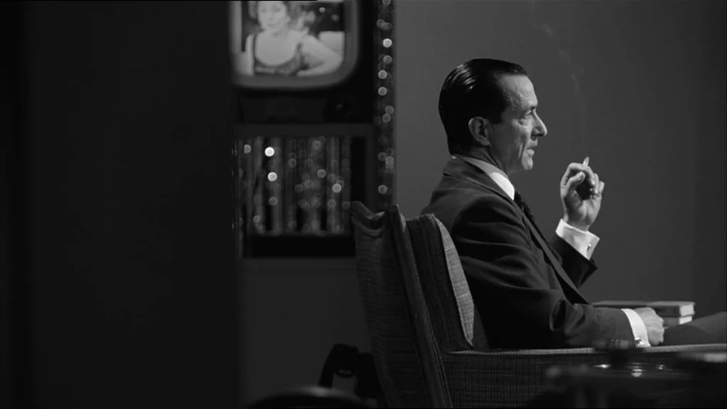 Good Night And Good Luck (2005) movie still: medium — A dark living room features a man sitting in a plush armchair facing right. He holds a lit…