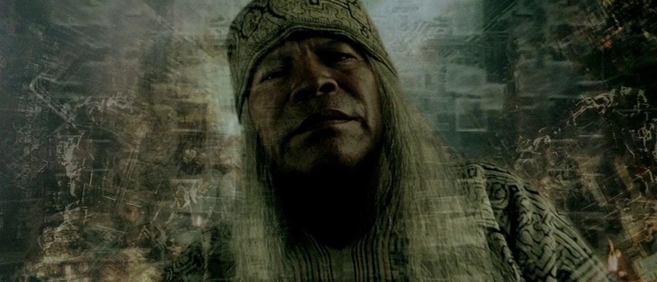 Renegade (2004) movie still: closeup — An older man wearing a patterned textile headpiece and shirt stares into the camera lens. Layered digital…