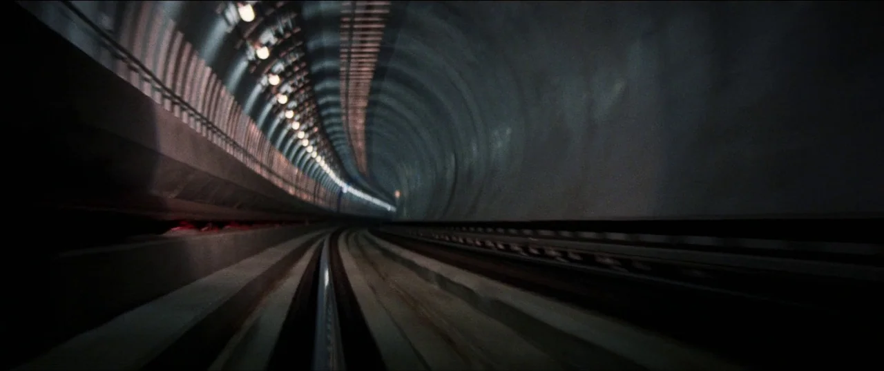 Speed (1994) movie still: establishing — A dark, curved underground train tunnel is constructed from smooth gray concrete segments. Rows of recessed…
