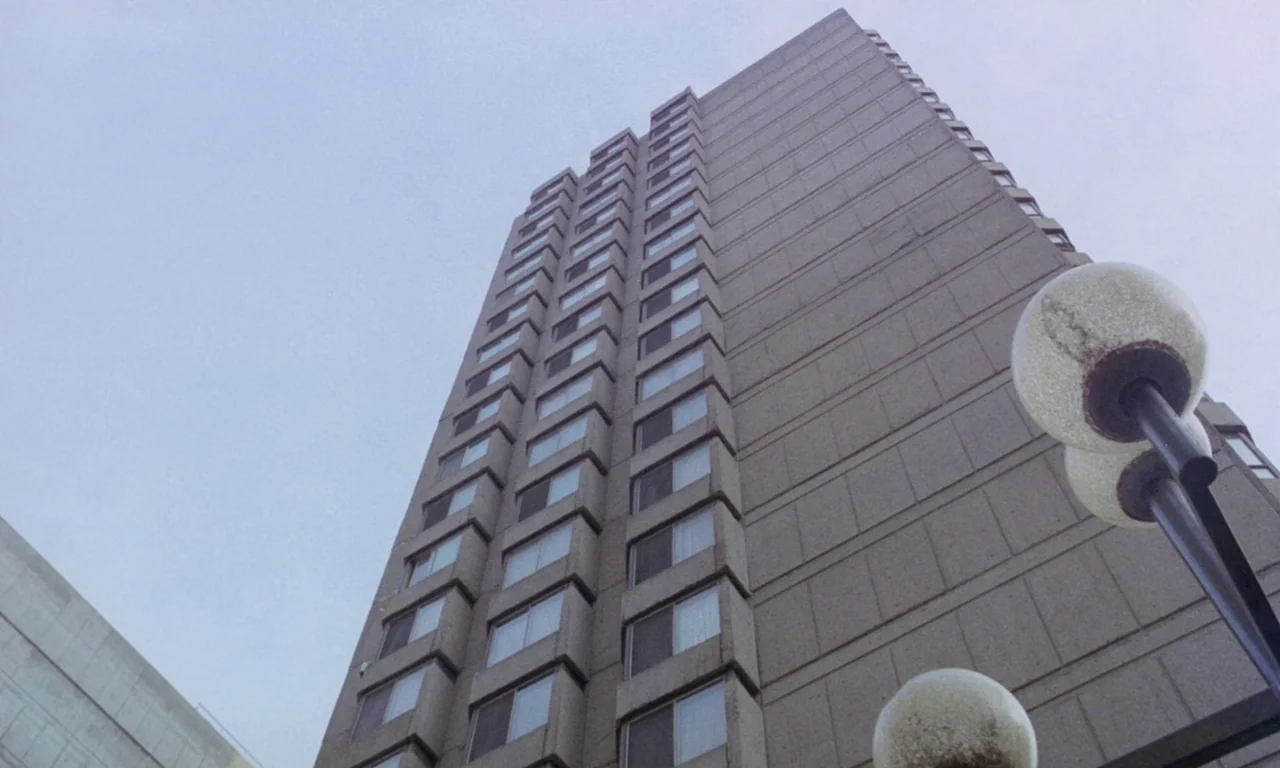 Rabid (1977) movie still: establishing — A tall, brutalist-style concrete apartment tower reaches upward against a pale overcast sky. Several…