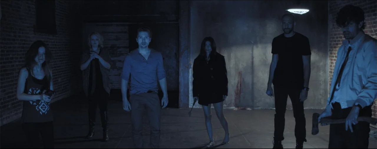El Último Juego (2025) movie still: fullbody — A spacious industrial area features raw brick walls and a dark concrete floor. Five adults stand…