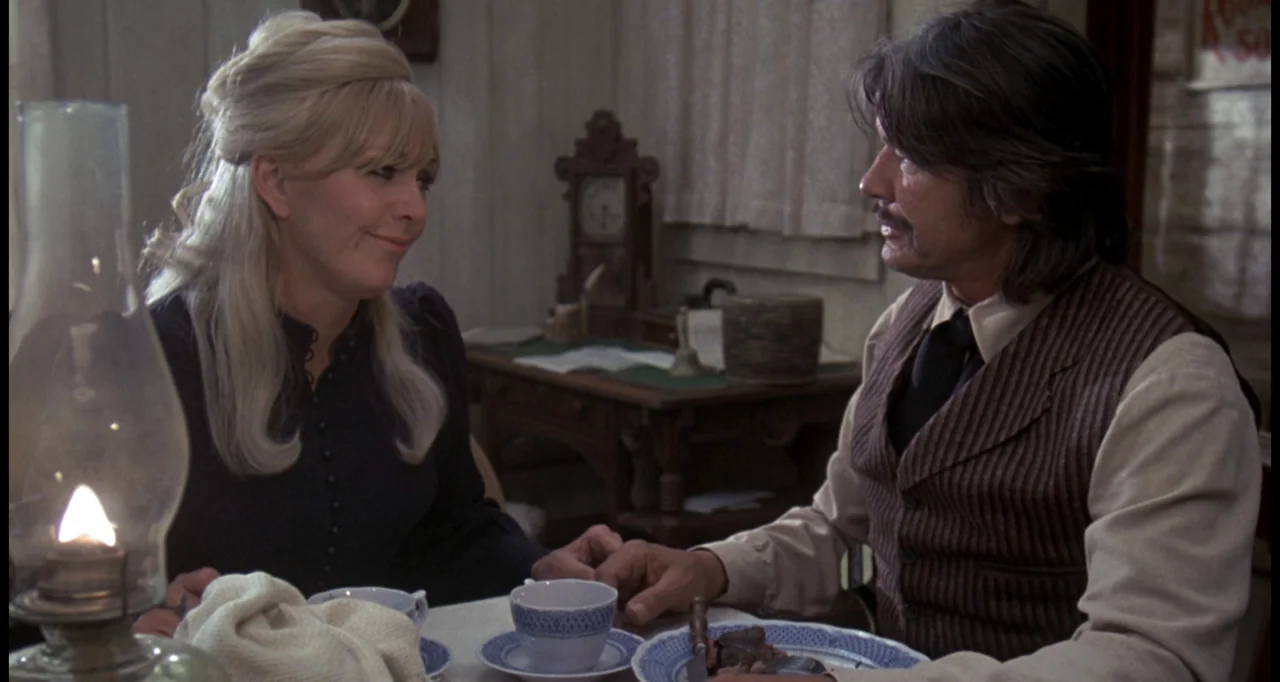 The White Buffalo (1977) movie still: medium — An interior space with wooden walls features a table set for a meal. A man and a woman sit across…