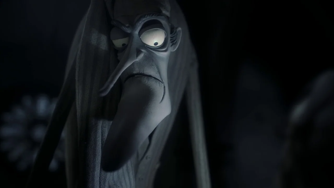 The Corpse Bride (2005) movie still: closeup — An animated, elongated figure with sharp facial features emerges from a deep black background. The…