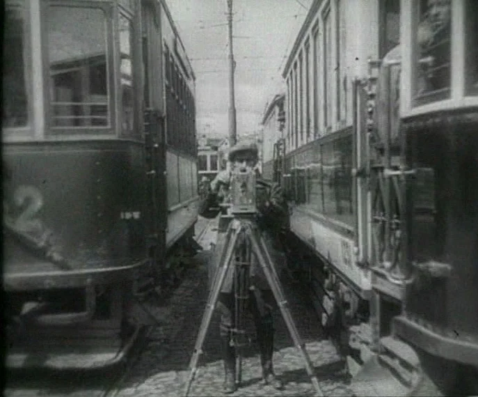 Man with a Movie Camera (1929) movie still: fullbody — A man stands on cobblestones between two parallel streetcars using a vintage film camera. The…