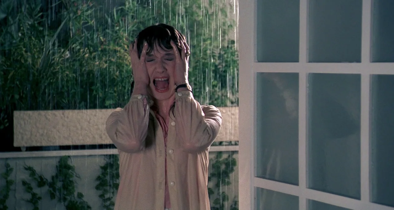 Tenebre (1982) movie still: medium — A woman stands in a rainy yard at night outside a house with glass doors. The woman is screaming loudly while…