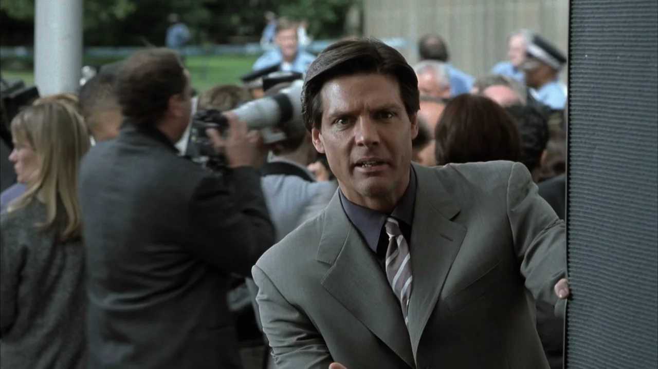 John Q (2002) movie still: medium — A man stands behind a dark partition surrounded by a crowd of press photographers. He leans toward the camera…