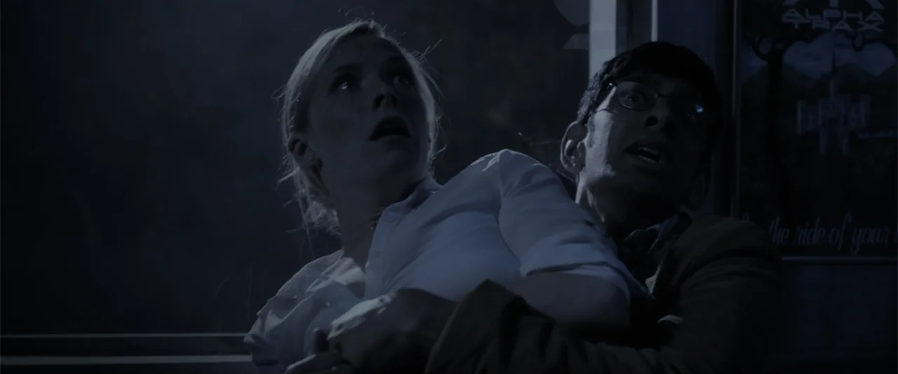 Howl (2015) movie still: medium — A dimly lit subway car interior featuring a dark wall with a poster visible. A woman is being held by a man as…