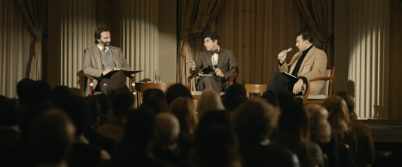 The Case for Christ (2017) movie still: wide — A wood-paneled conference hall stage features three men seated in armchairs before an audience. The…