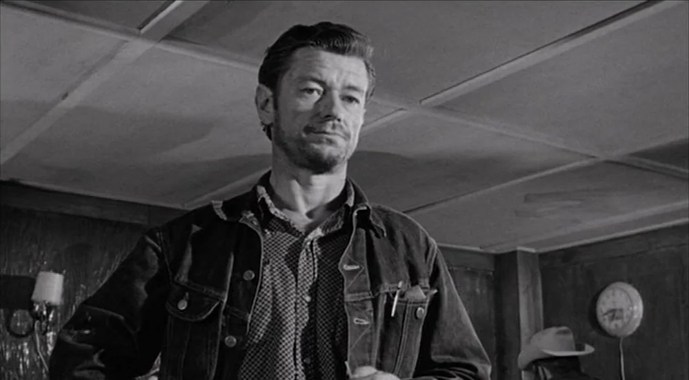 The Last Picture Show (1971) movie still: medium — A dimly lit room with wood-paneled walls and a ceiling featuring recessed square panels. A man…