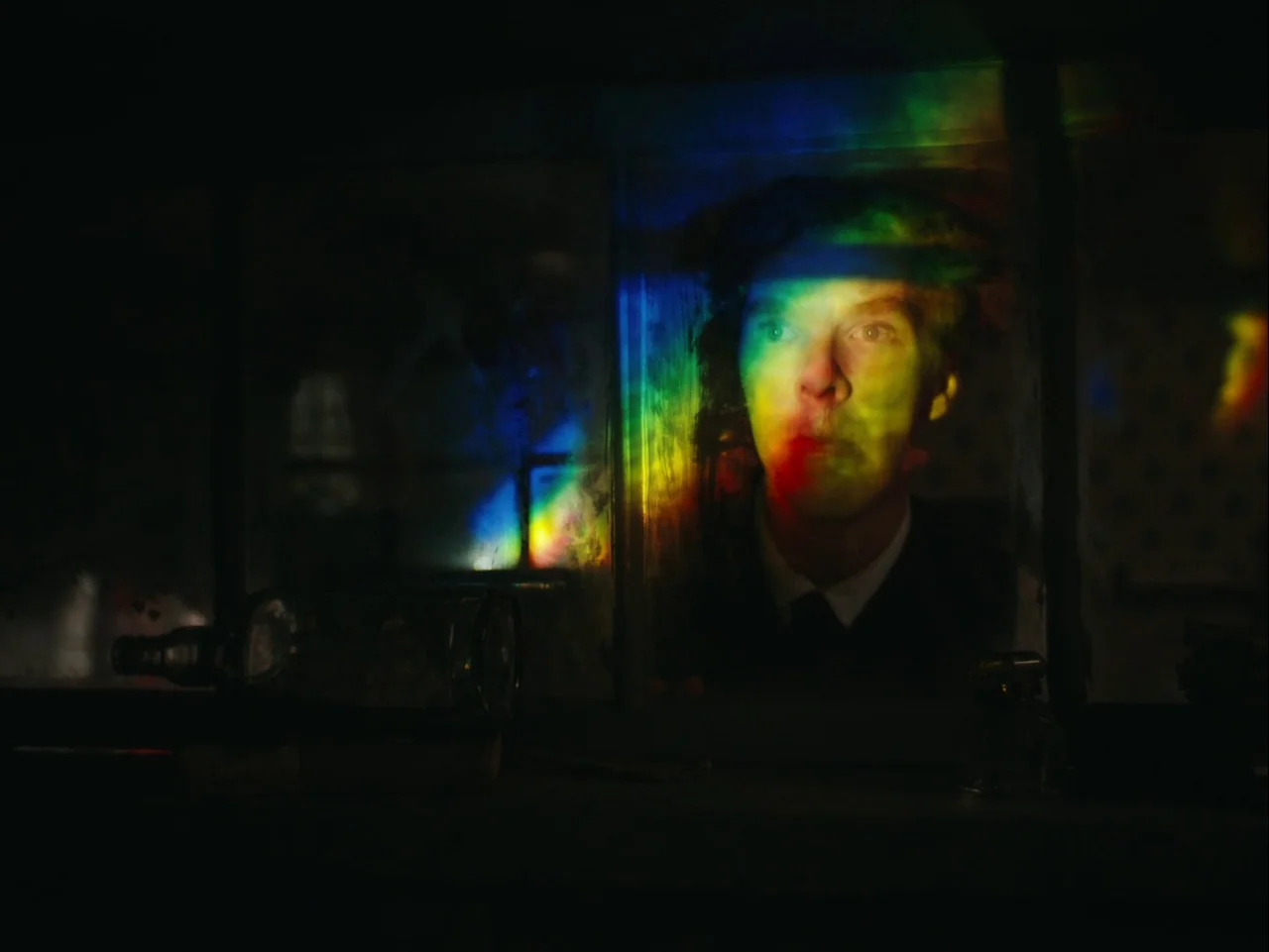 The Electrical Life of Louis Wain (2021) movie still: closeup — A dark space features a man visible through a glass partition. Vibrant multicolored…