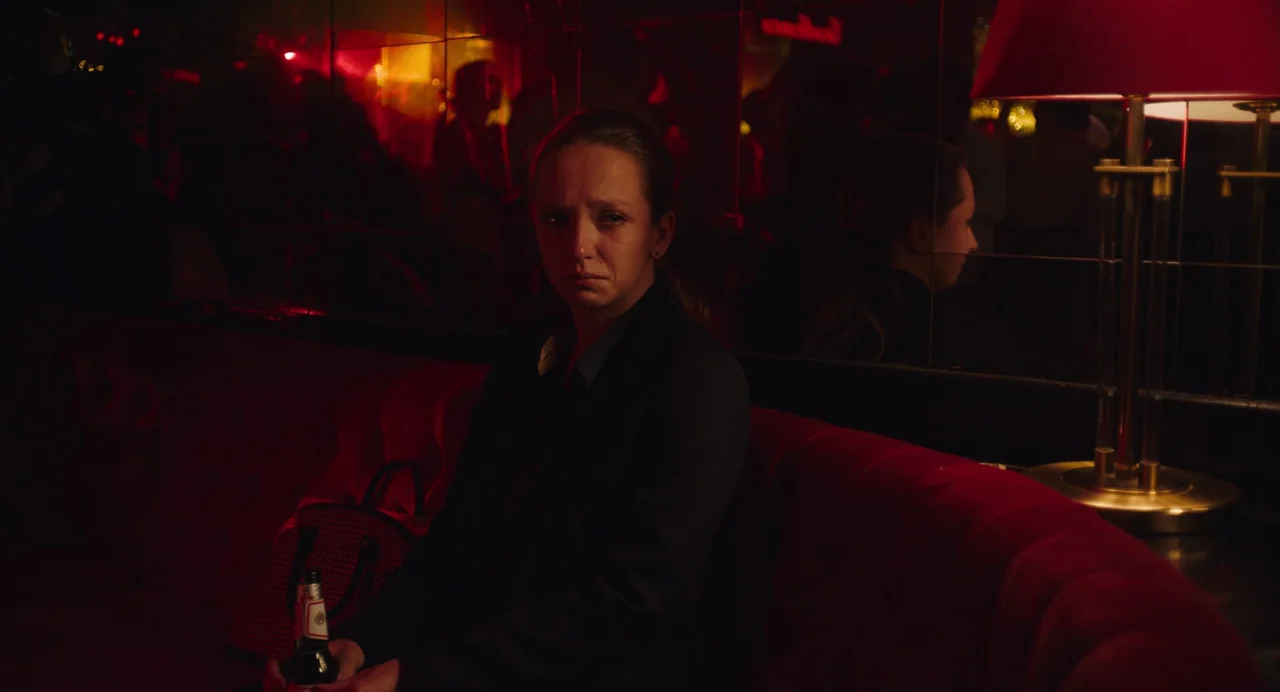 She Said (2022) movie still: medium — A woman sits in a dark booth with mirrored walls and red velvet furniture. She rests on the seat with a beer…