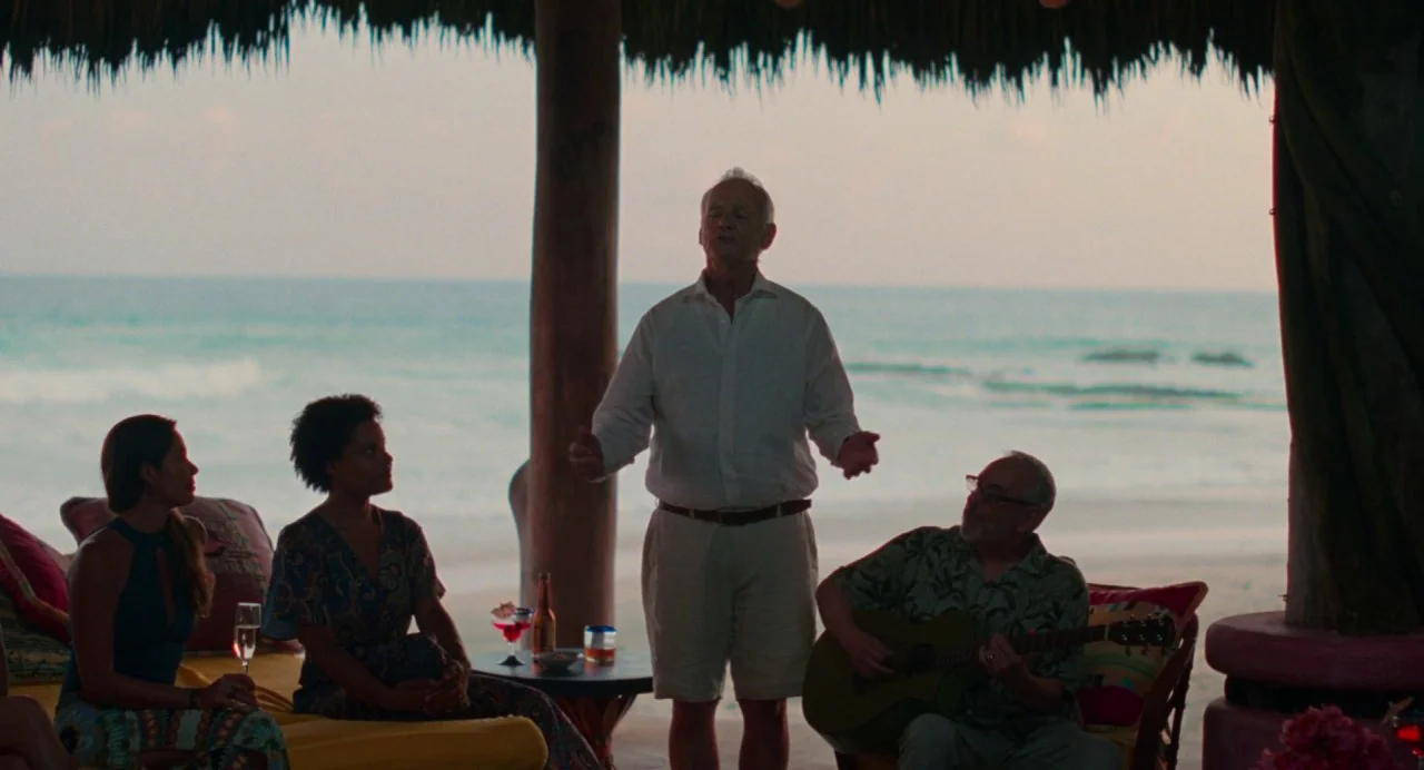 On The Rocks (2020) movie still: fullbody — A beachside structure with a thatched roof sits above a quiet ocean shoreline at dusk. An elderly man…