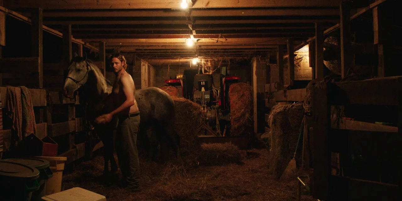 The Death of Dick Long (2019) movie still: fullbody — A dimly lit wooden stable contains hay bales, a tractor, and a grey horse. A shirtless man…