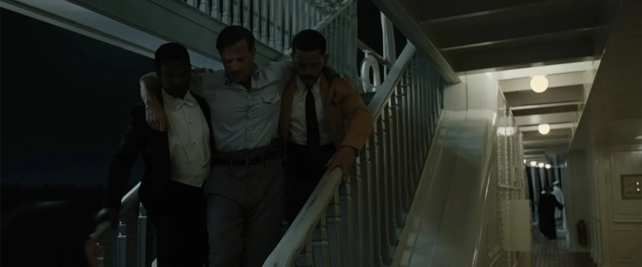 Death on the Nile (2022) movie still: fullbody — A nighttime outdoor passenger ship staircase with white railings and warm deck lights. Three men…