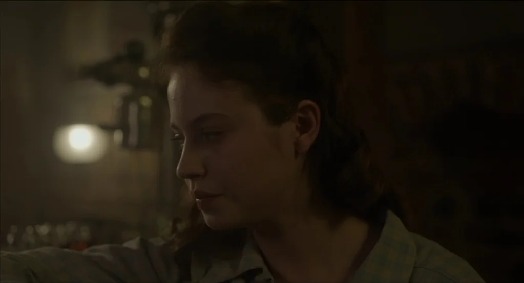 The Innocents (2016) movie still: closeup — A dimly lit interior room at night features a soft glowing practical lamp. A young woman with dark hair…