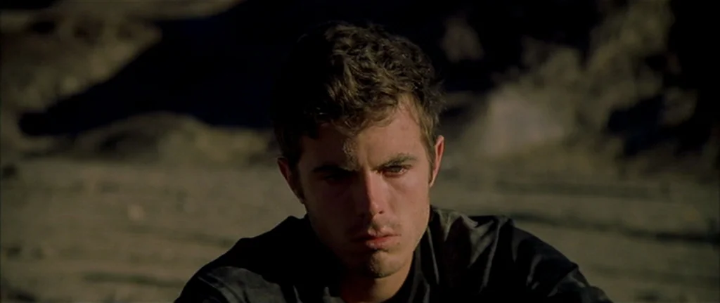 Gerry (2002) movie still: closeup — A man sits in an arid, rocky, and sun-drenched outdoor environment. He gazes downward with a still expression.