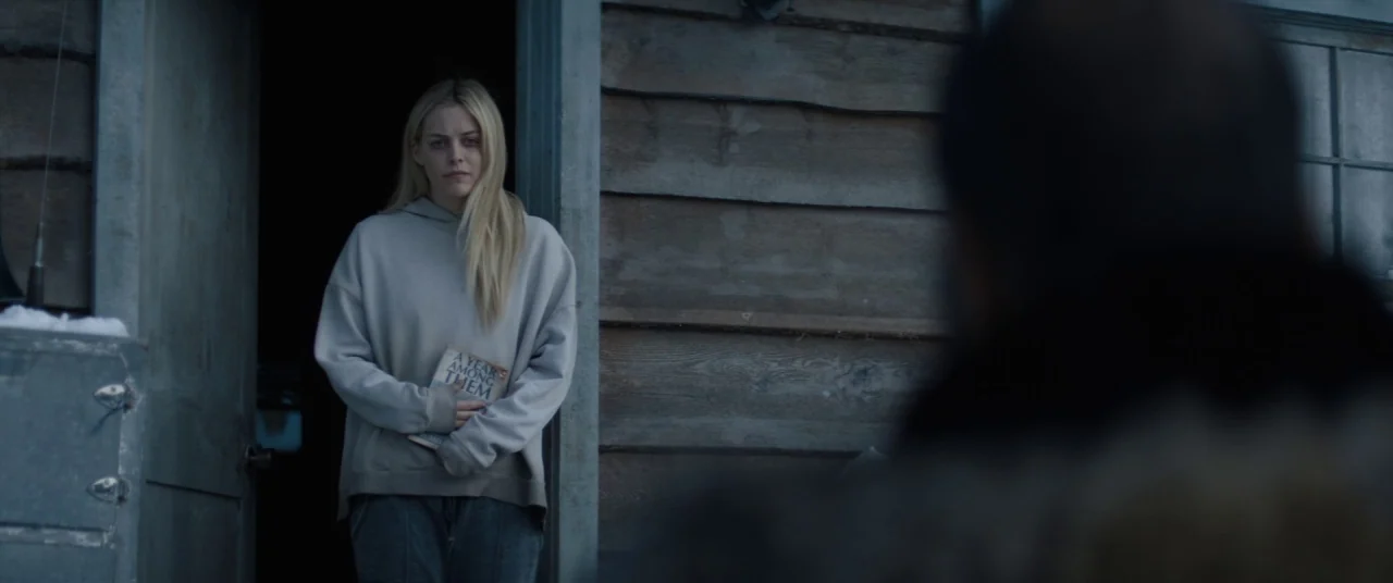 Hold The Dark (2018) movie still: medium — A woman stands in the doorway of a weathered wooden cabin in an overcast climate. She clutches a book…