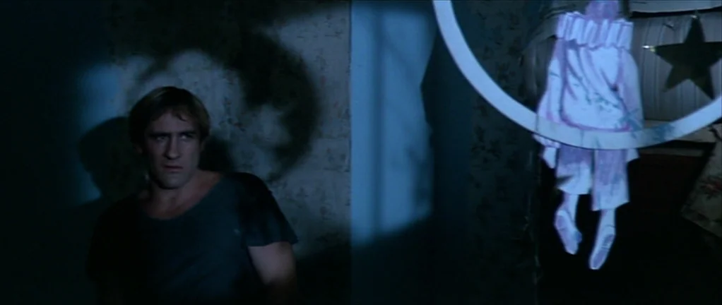 The Moon in the Gutter (1983) movie still: medium — A dark, dimly lit room contains patterned wallpaper and a suspended mobile. A man sits at the…