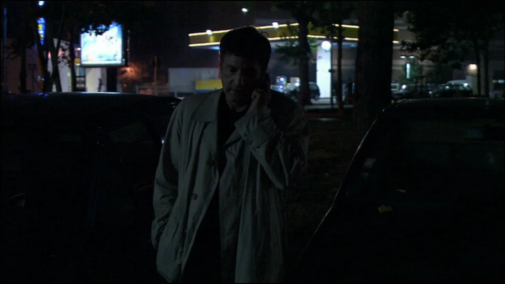 Caché (Hidden) (2005) movie still: medium — A man stands between parked cars in a dimly lit, nocturnal outdoor space. He holds a phone to his ear…