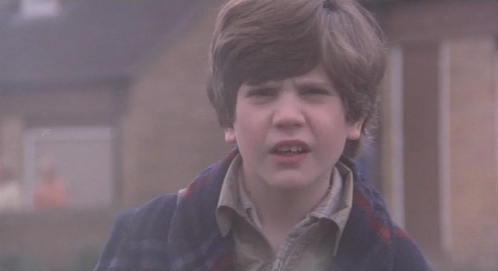 Time Bandits (1981) movie still: closeup — A suburban residential area is obscured by a thick layer of grey fog. A young boy stands in the…