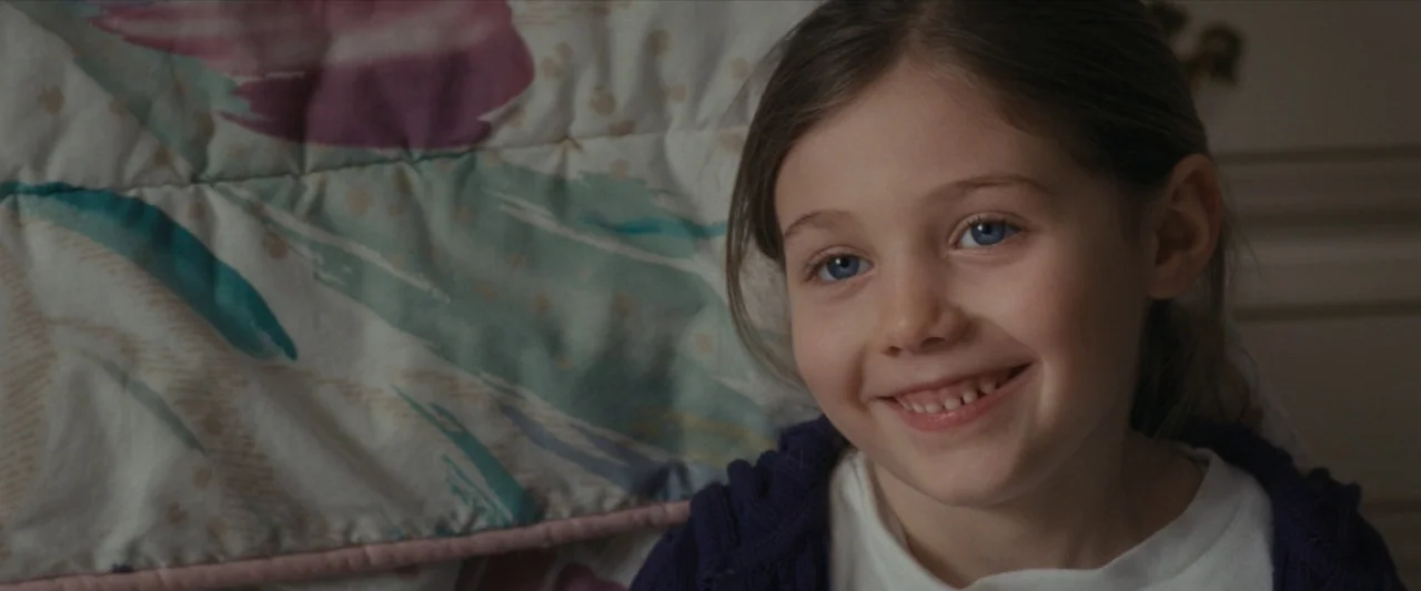 For Ellen (2012) movie still: closeup — A young girl sits in front of a patterned floral quilt. She looks toward the side with a bright, open smile.