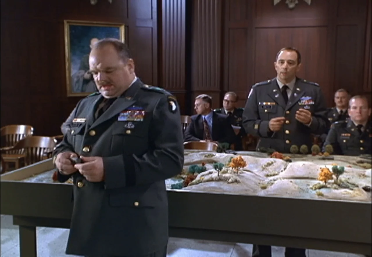 The Pentagon Wars (1998) movie still: medium — A wood-paneled room contains a large table displaying a detailed topographic military model map. Two…