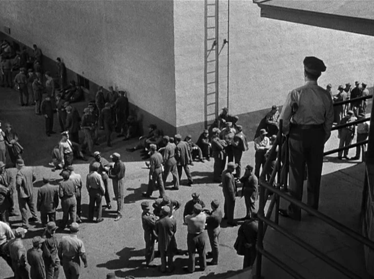 White Heat (1949) movie still: wide — A large concrete prison yard is filled with many men in work clothing. A lone uniformed guard stands on a…