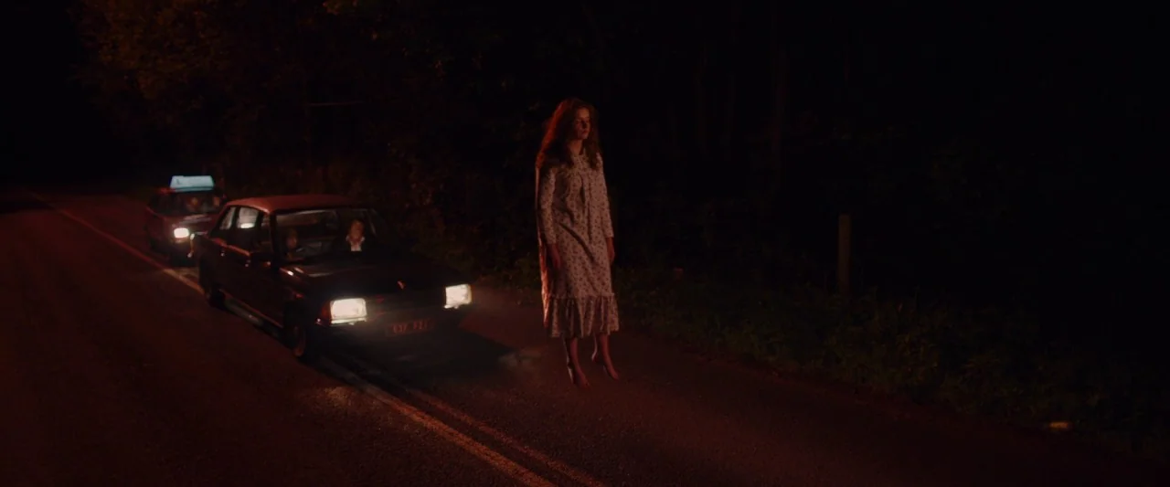 Extra Ordinary (2019) movie still: fullbody — A dark, unlit rural road at night bordered by trees. A young woman in a nightgown levitates inches…