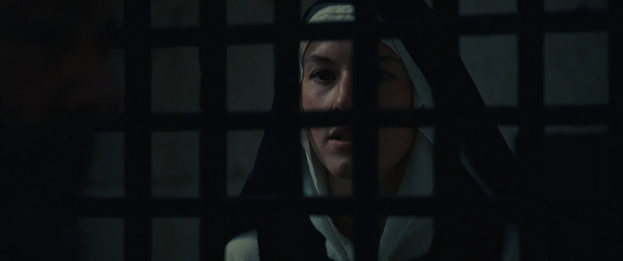 Benedetta (2021) movie still: closeup — A woman wearing a nun's habit stands behind a dark, rectangular metal lattice grate. She peers through the…