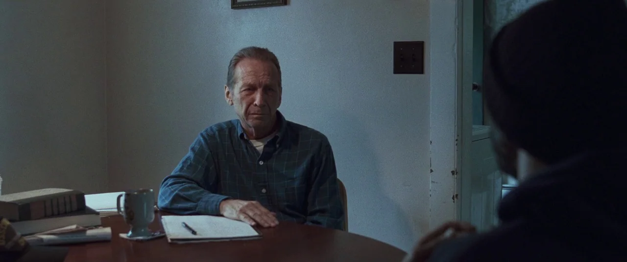 Sound of Metal (2019) movie still: medium — A spare room features a dark circular wooden table against a plain gray wall. An older man sits behind…