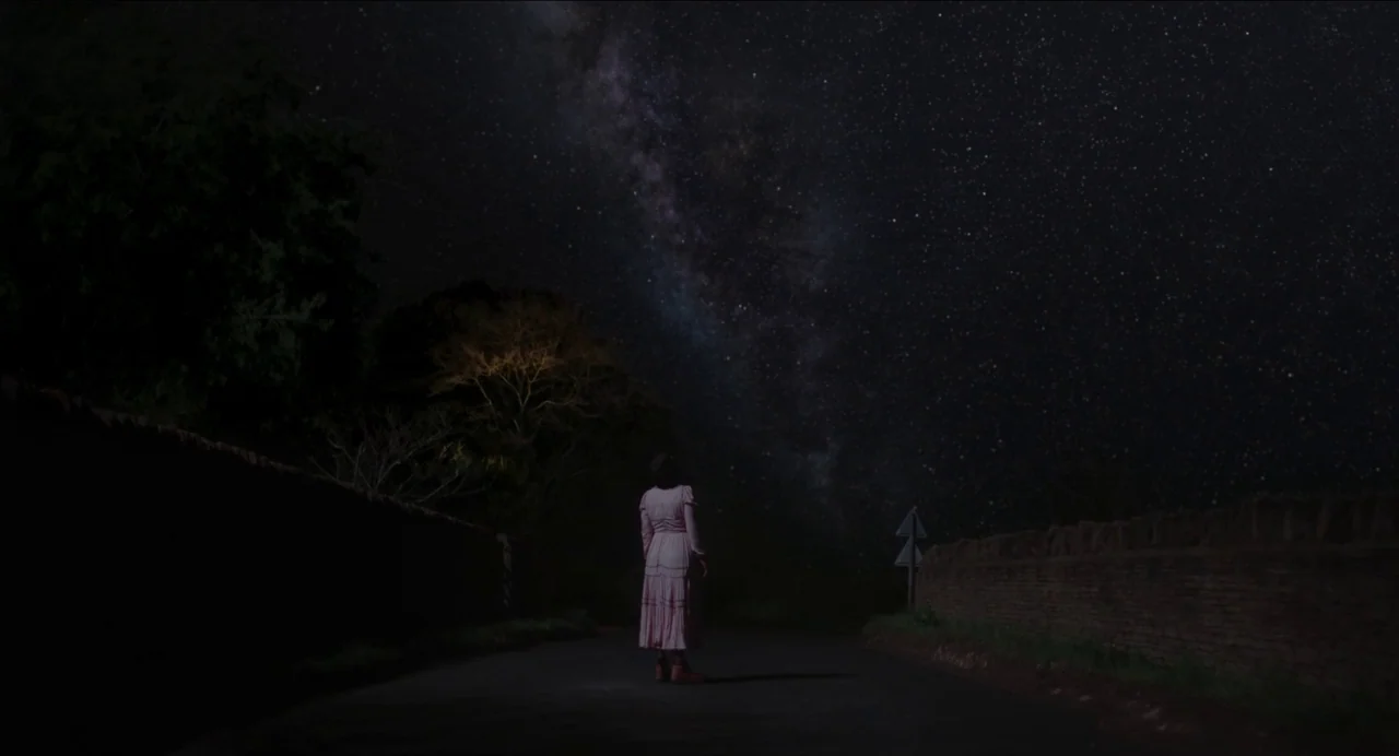 Men (2022) movie still: wide — A dark rural road stretches between stone walls beneath a star-filled night sky. A lone woman in a long dress stands…