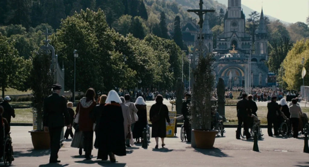 Lourdes movie still: wide — A wide paved plaza leads toward a large ornate stone cathedral in the distance. Many people walk in the same direction…