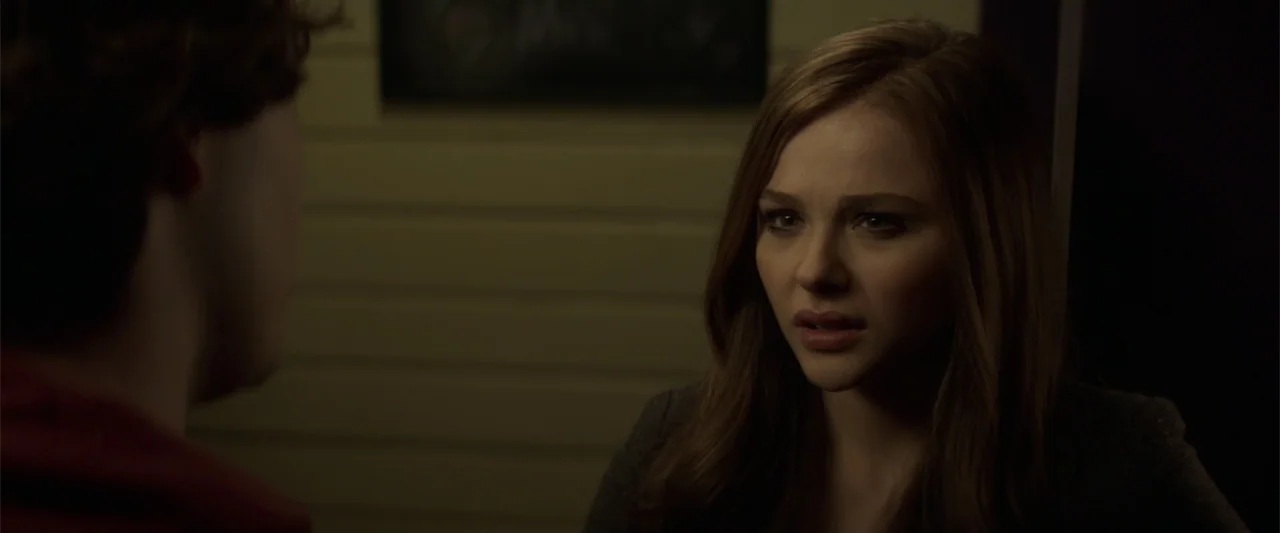 If I Stay (2014) movie still: medium — A dimly lit interior space with horizontal wall panels. A young woman with brown hair stares intently at…