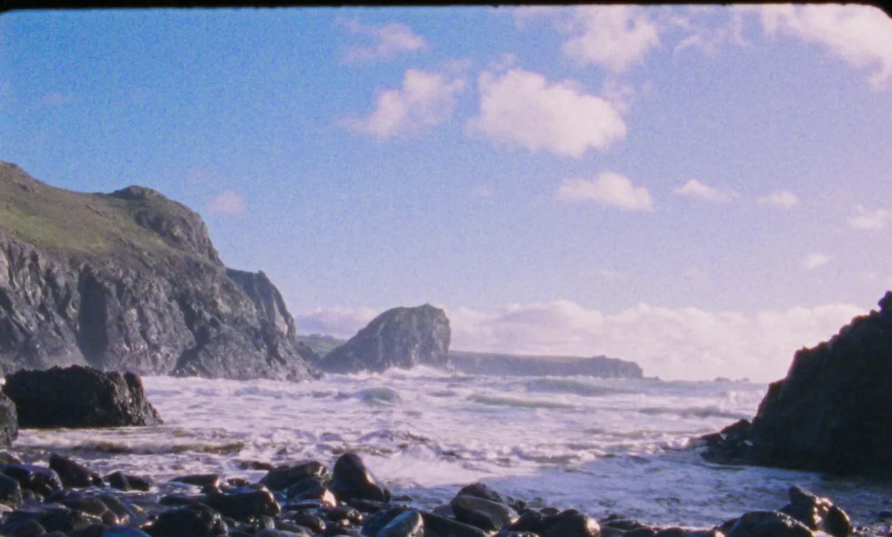 The Silent Twins (2022) movie still: wide — A rugged coastline featuring a rocky beach with dark stones and crashing ocean waves. Large dark cliffs…