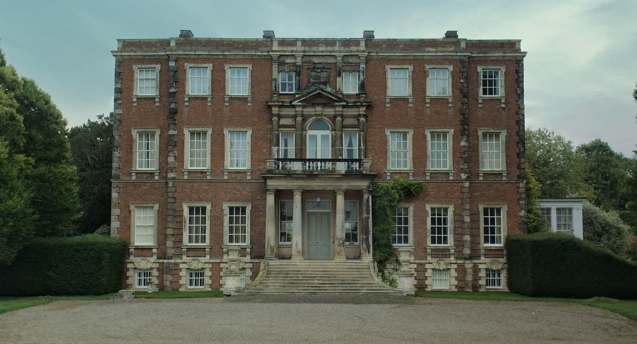 Flux Gourmet (2022) movie still: establishing — A symmetrical brick stately home features three stories and a central stone entryway. Ivy grows up…