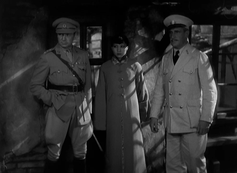 Shanghai Express (1932) movie still: medium — A dark indoor space with rough stone walls and a paneled window frame. Two men in official uniforms…