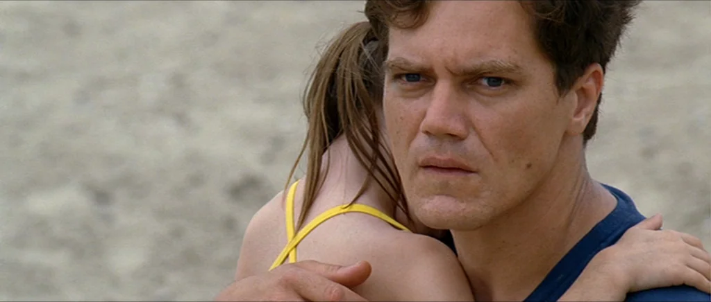 Take Shelter (2011) movie still: medium — A man and a young girl embrace on a sandy beach during the day. The man looks ahead while the girl faces…
