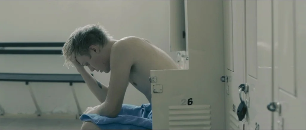 Lasting (Nieulotne) (2013) movie still: medium — A locker room with beige metal lockers features a single wooden bench. A young man sits shirtless…