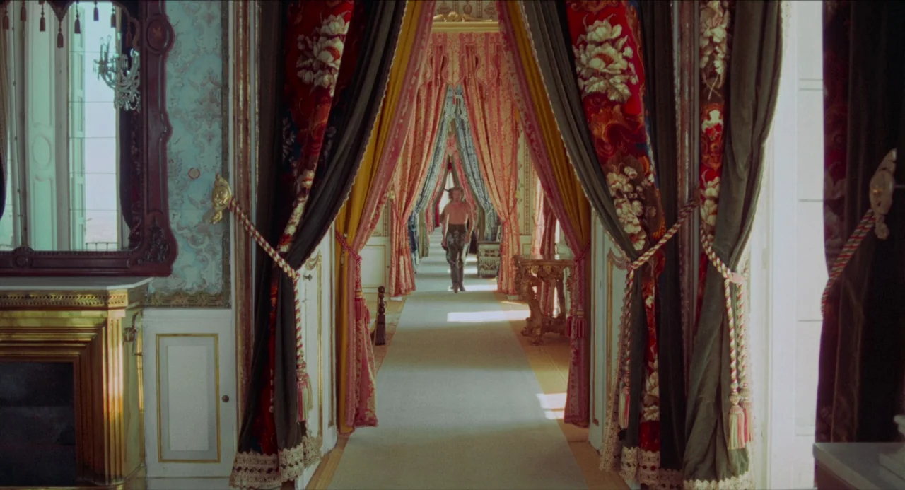 The Three Musketeers (1973) movie still: wide — A long, opulent hallway is lined with heavy, patterned velvet curtains and ornate molding. A lone…