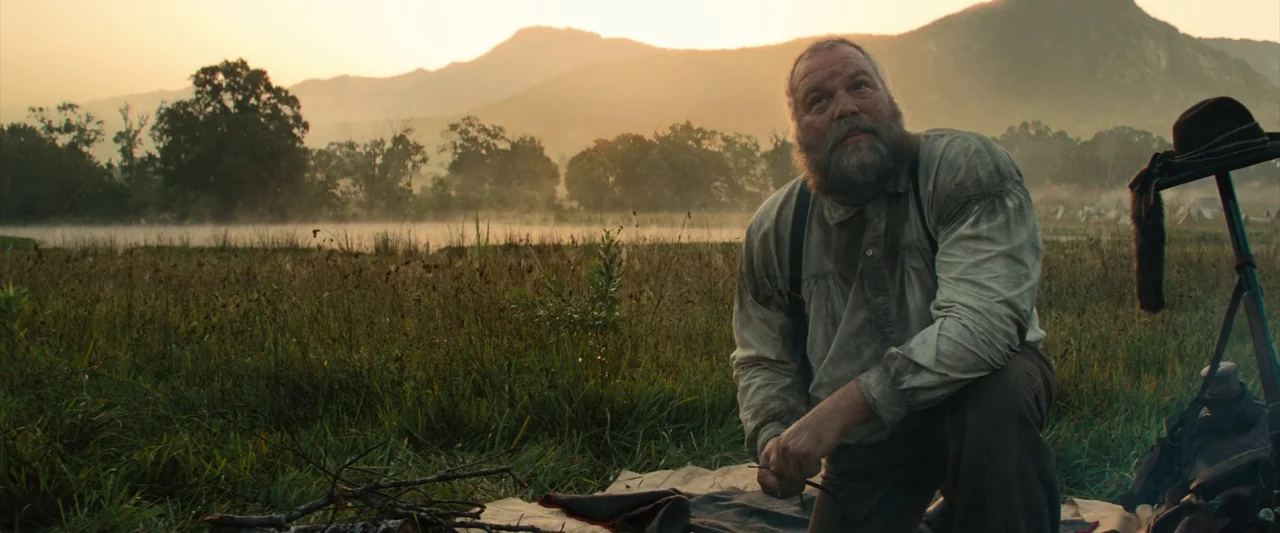 The Magnificent Seven (2016) (2016) movie still: medium — A grassy meadow at dawn features a thin layer of morning mist. A man kneels on a blanket…